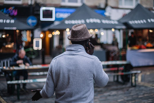 A man on the phone in front of street food stalls strung with lights at dusk.