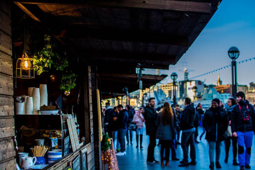 A wooden riverside food stall lit by a lantern at dusk, with a crowd walking past.